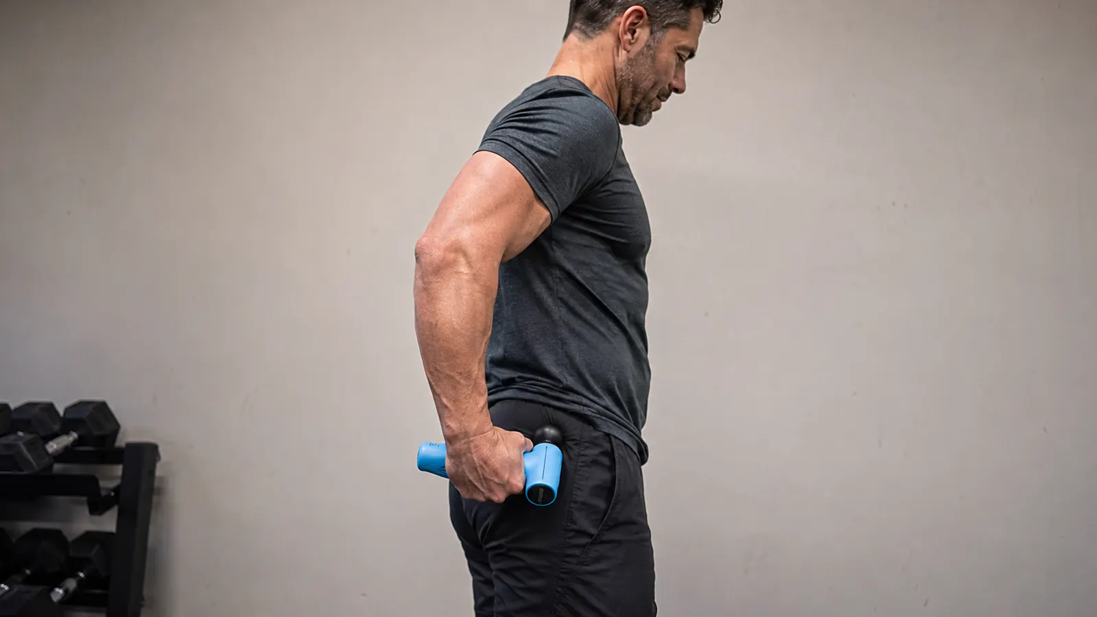 Man using the BOB AND BRAD C2 massage gun on his lower glute area while standing against a plain gym wall, flat attachment visible