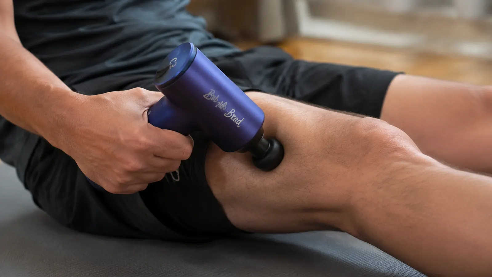 Hand pressing a percussion massage gun against a quad muscle on a foam mat