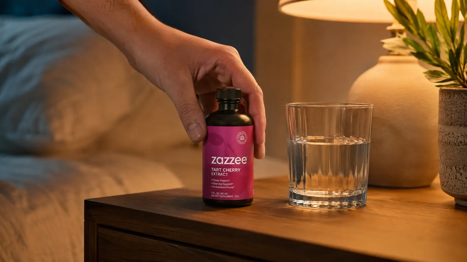 Hand holding a Zazzee tart cherry extract capsule bottle next to a glass of water on a nightstand